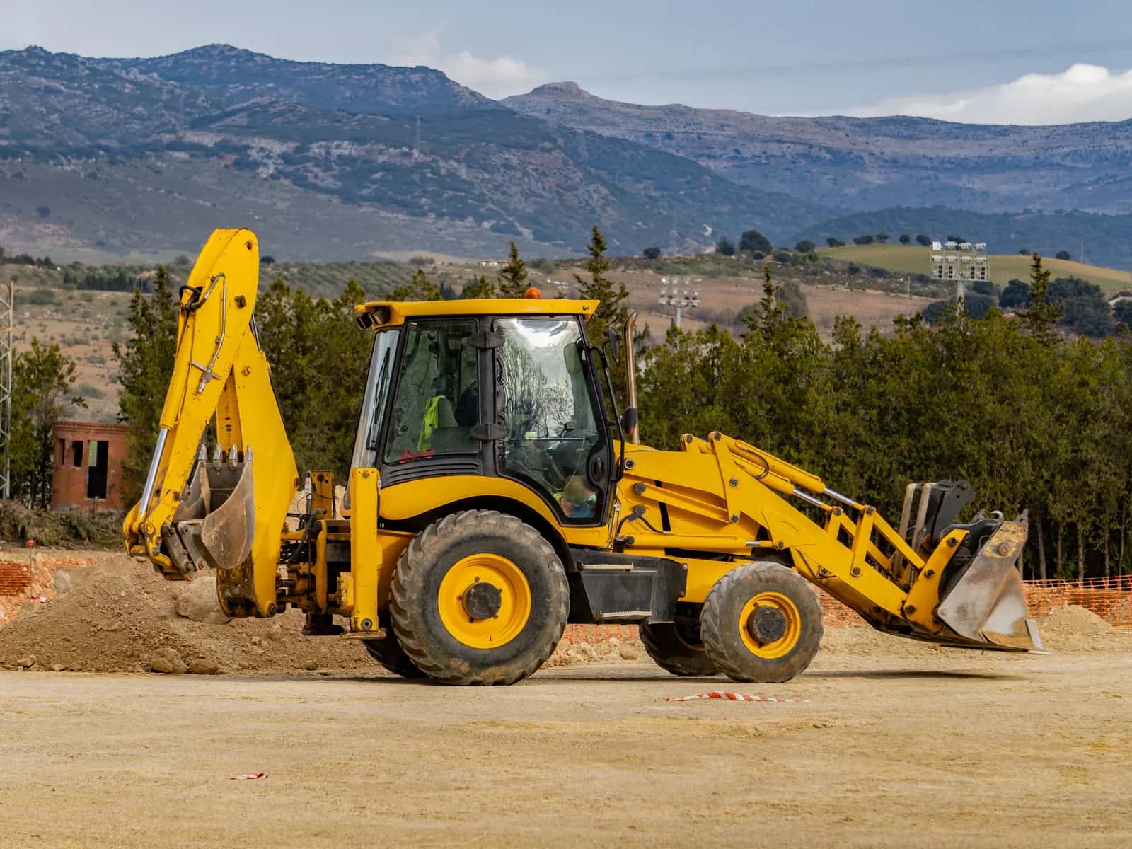 Backhoe loader kuning dengan bucket muat di depan dan lengan gali di belakang, di lahan proyek